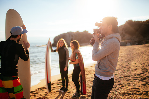 Young man holding video camera, having a photo shoot with his friends at the beach, while they are preparing to surf