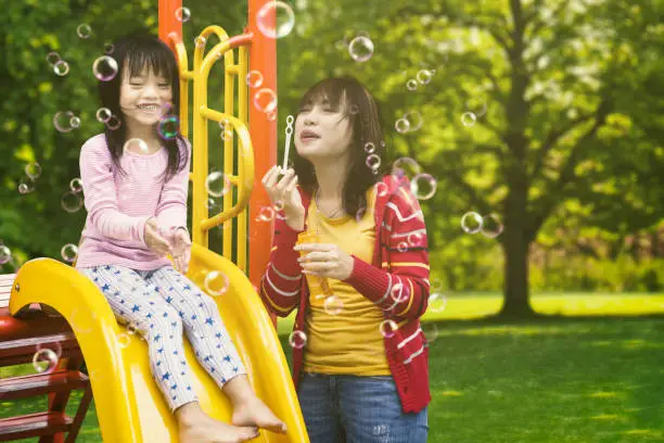 Portrait of beautiful mother and cute child playing soap bubbles while sitting on the playground
