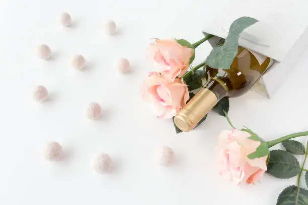 White wine bottle and three pink roses in a silver glitter gift bag on white background surrounded by champagne truffles.