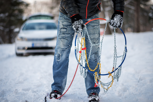 Uknown person is holding the snow chains before mounting to a car tire. White vehicle is in the background.