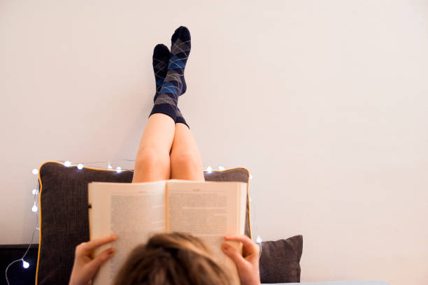 winter, coziness, leisure and people concept - close up of young woman with Beautiful female legs on the pillow reading book in bed at home