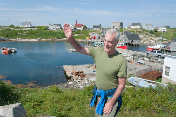 Handsome Senior Man Waving to Friend While Standing on Bluff Overlooking Fishing Village stock photo