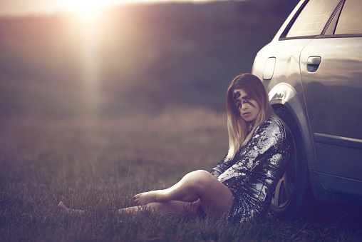 warrior woman with dirt on her face sitting near car and thinking far away. photo taken in the sunset in nature. wearing shiny sequin dress.