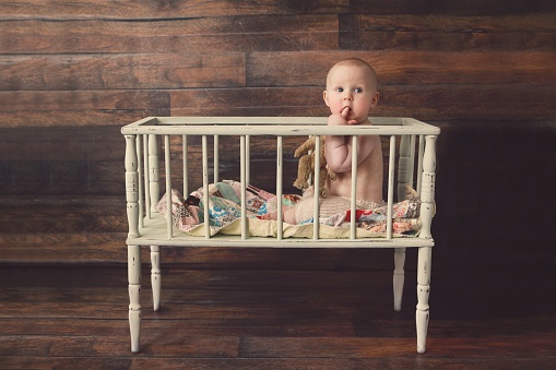 A baby boy, six months old, sits in a vintage wooden crib.