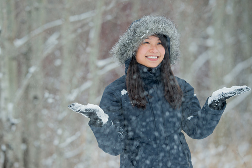 A beautiful Asian woman catches snow on a cold winter day while wearing a fur lined coat.