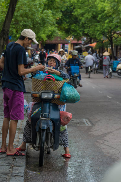Buying vegetables Central Hoi An Street. A woman, sitting on her moped, pays the purchase of vegetables she has just made, to the man standing with her. andar en bicicleta stock pictures, royalty-free photos & images