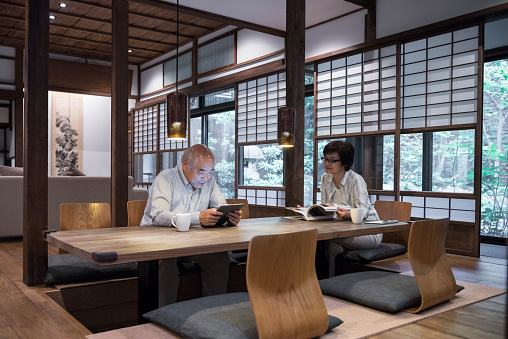 Senior man and mature woman using digital devices in traditional Japanese home, sitting at wooden table