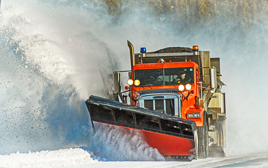 Orange snowplow makes its way down the highway, clearing the snow as it goes.  Picture was taken in Interior Alaska on a bright day in winter.