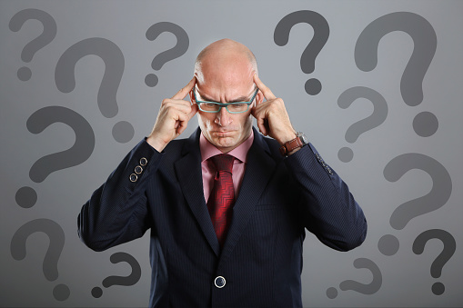 Portrait of a mid adult male businessman thinking with question marks around him. About 35 years old, Caucasian male with shaved head and glasses.