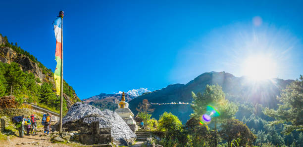 Bright high altitude sun shining on trekkers and porters as they walk through a Sherpa village past prayer flags, mani stone and Buddhist stupa high on the Everest Base Camp trail in the Himalayan mountains of Nepal.