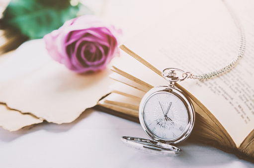 Close up of antique silver pocket watch and opened book with rose flower and old letter papers on white table background with vintage light