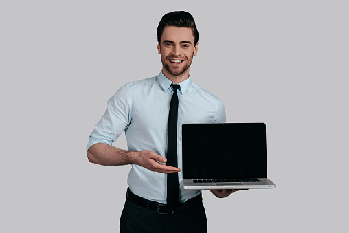 Good looking young man in white shirt and tie pointing copy space on his laptop and smiling while standing against grey background