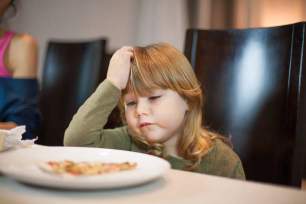 child does not want pizza gesture at home Three years old blonde child looking at pizza piece in her dish with expression she does not like it, sitting in dark brown chair, in table with beige tablecloth at home, next to mother fat kid eating pizza stock pictures, royalty-free photos & images