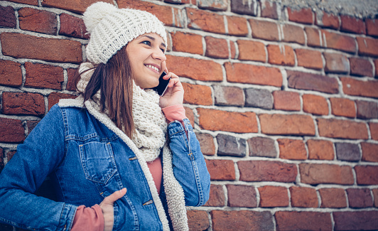 Beautiful young brunette woman, standing by the brick wall, enjoying the cold air outdoors, using her telephone.