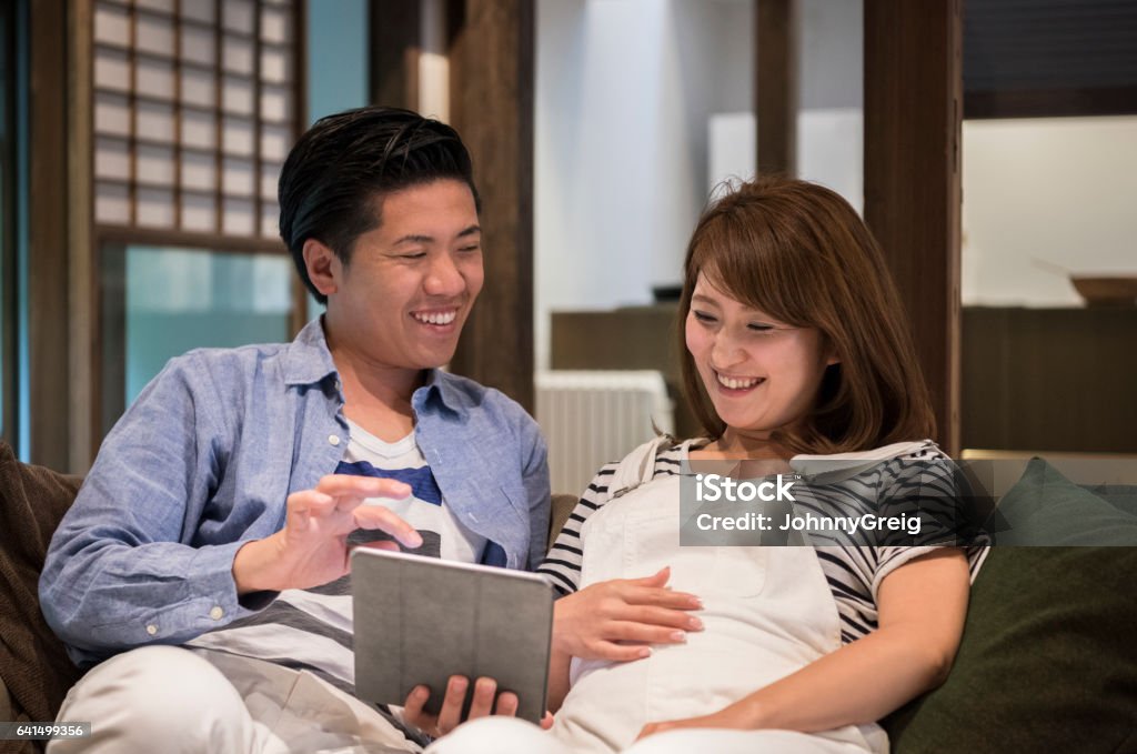 Japanese couple using tablet, pregnant woman with hand on tummy Happy mid adult couple looking at tablet and smiling, woman touching belly, man holding tablet, laughing Digital Tablet Stock Photo