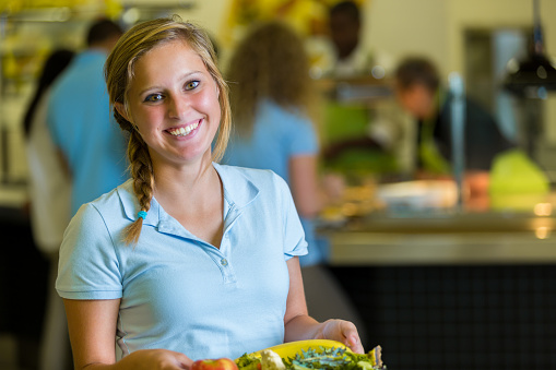 Confident Caucasian teenage girl holds tray of healthy food in school cafeteria. Students are in line behind her. She is smiling in the camera.