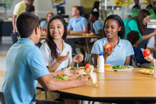 Diverse teenage friends eat lunch in school cafeteria Male and female teenagers enjoy eating lunch together in the school cafeteria. They are wearing school uniforms. cafeteria stock pictures, royalty-free photos & images