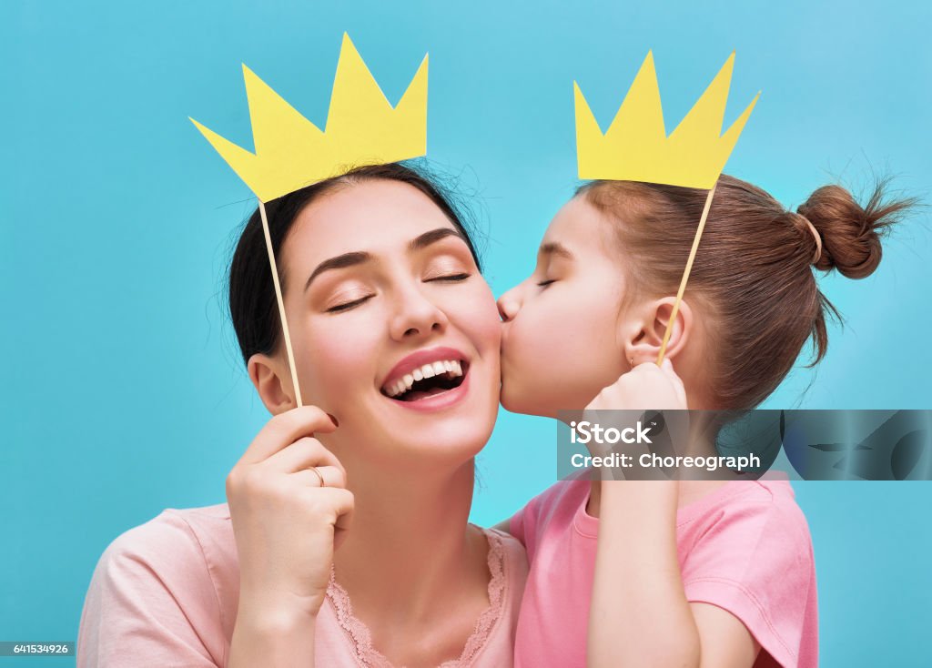 Happy loving family Funny family on a background of bright blue wall. Mother and her daughter girl with a paper accessories. Mom and child are holding paper crown on stick. Mother Stock Photo