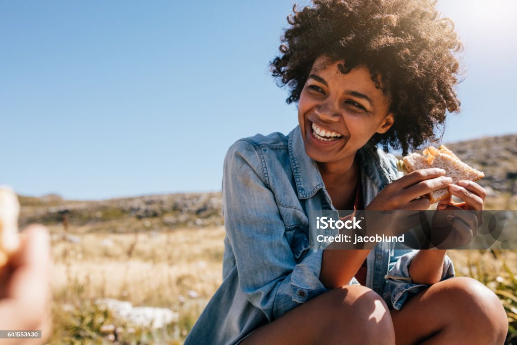 Happy young woman taking break during country hike. Happy young african woman eating sandwich and smiling. Taking break during country hike. Eating Stock Photo