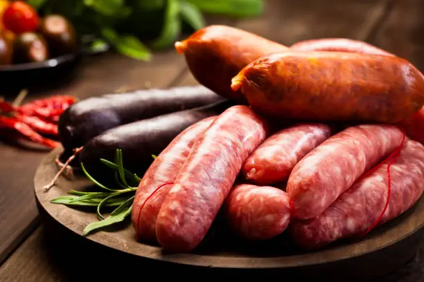 Wooden plate of raw meat sausages variation shot on dark wood table. Some herbs and spices are out of focus at the background. Predominant colors are red and brown. DSRL studio still life taken with Canon EOS 5D Mk II and Canon EF 100mm f/2.8L Macro IS USM