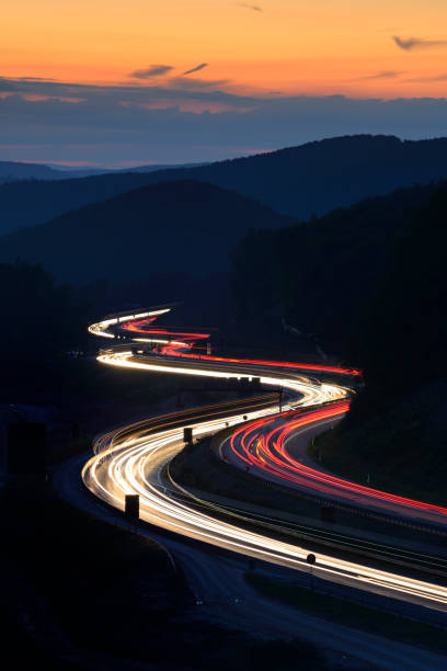 lunga esposizione di luci per auto sull'autostrada che serpeggiano tra le colline al tramonto - curve road winding road street foto e immagini stock