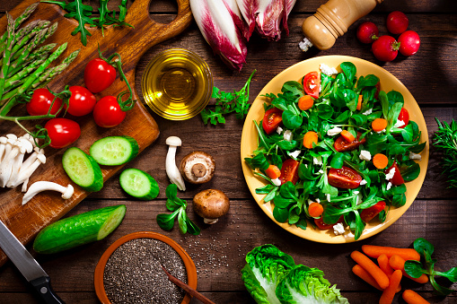 Top view of a rustic wood table filled with ingredients for preparation of a healthy salad with chia seeds. The ingredients included for the preparation are chia seeds, lettuce, cherry tomato, carrot, cucumber and goat cheese. A served salad plate is at the right of the frame while a wooden cutting board with some vegetables is at the left. The salad is served on a yellow plate. Some vegetables for salad preparation are scattered on the table. DSRL studio photo taken with Canon EOS 5D Mk II and Canon EF 100mm f/2.8L Macro IS USM