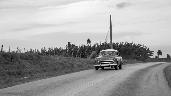 Cienfuegos, Cuba - March 21, 2016: Black and white photograph of a classic black and white  car driving on a Cuban Highway. There are  some bushes around the road and the sky has some clouds.