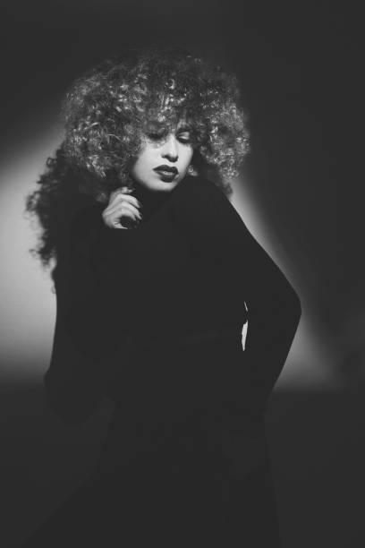 monochrome shot of curly hair woman in black dress posing in studio, looking down, seductive expression.
