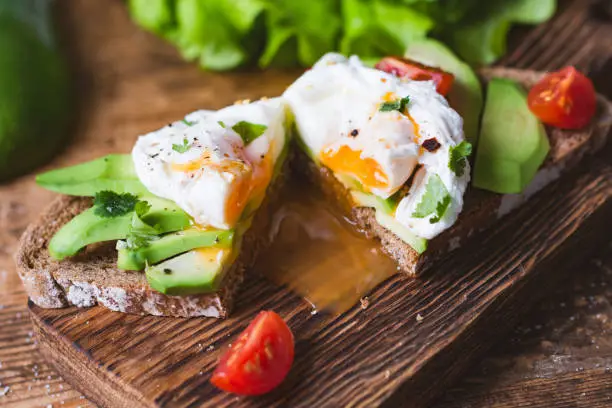 Poached egg, avocado snack toast sandwich on wooden cutting board. Close up view, selective focus