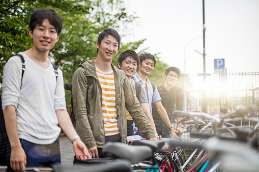 Portrait of happy university students standing by bicycles. Male friends smiling together on sunny day. All are at parking lot.