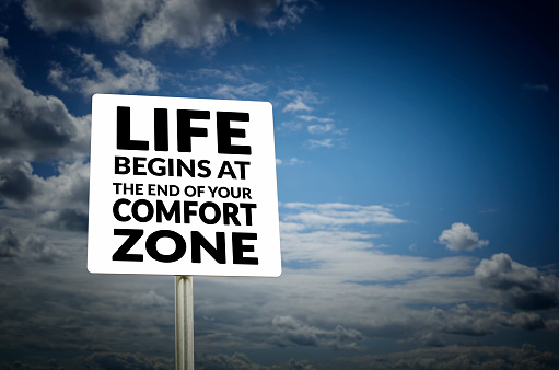 Life begins at the end of your comfort zone road sign with blue sky and clouds background