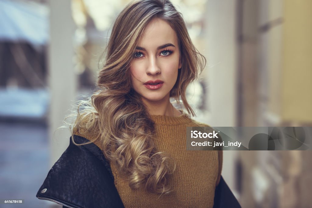Blonde woman in urban background. Blonde woman in urban background. Beautiful young girl wearing black leather jacket and mini skirt standing in the street. Pretty russian female with long wavy hair hairstyle and blue eyes. Women Stock Photo