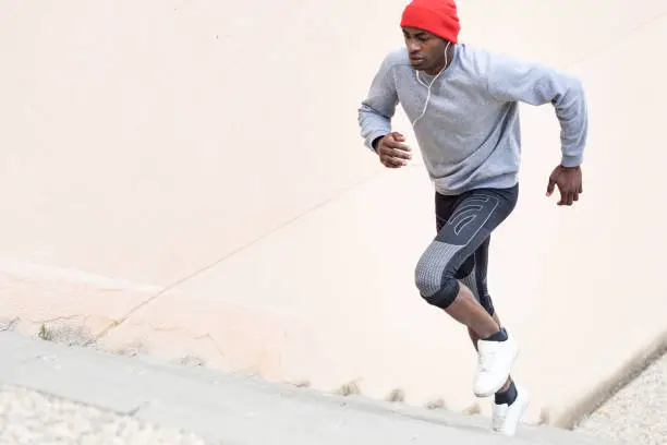 Black man running upstairs outdoors listening to music with white headphones. Young male exercising with city scape at the background.