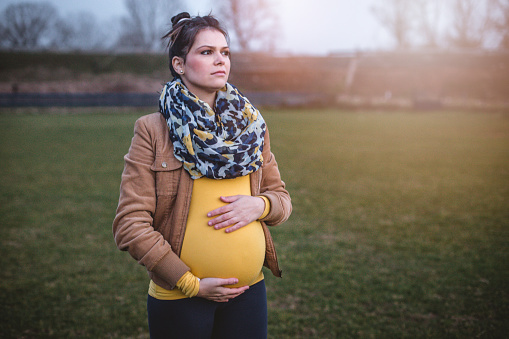 Alone, pregnant woman with her baby. Feeling worried and afraid. Holding her belly. She is outside in a park on a grass field