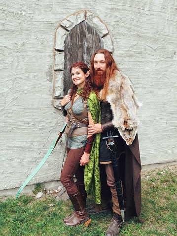 Two male Viking warrior men in authentic garb and armour travel in the forest with a female wood elf fairy fantasy character.Viking Norse Medieval Mythology, Warrior, Female Elf Relationship. Viking warrior and elf couple standing in front of castle facade.