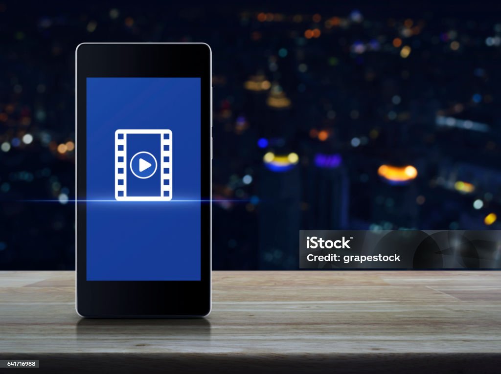 Cinema online concept Play button with movie icon on modern smart phone screen on wooden table over blur colorful night light city tower, Cinema online concept Vitality Stock Photo