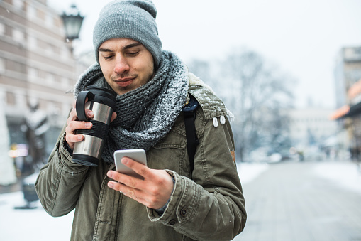 Young man checking his smart mobile phone outdoors while walking down the street on winter day, holding a travel mug with hot coffee.