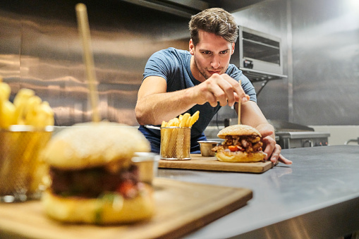 Chef preparing burger on wooden plate. Handsome professional is wearing casuals. He is working in restaurant.