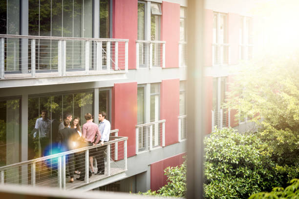 A photo of business people discussing at balcony. Coworkers are seen through glass window. They are standing by railing of office building.