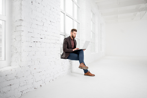 Elegant businessman sitting on the window with laptop in the white office interior
