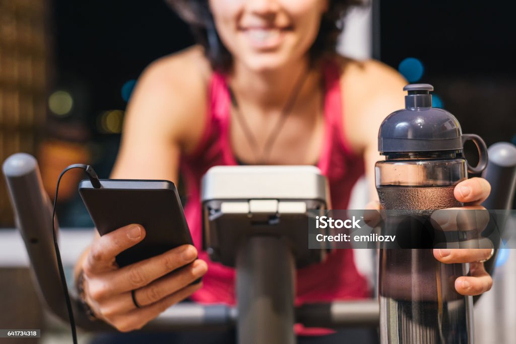 Healthy fit smiling woman training at home on exercise bike Healthy fit smiling woman training at home on exercise static bike during workout holding phone and bottle of water while listening music with earphones for motivation. Female health weekly habits app Bicycle Stock Photo