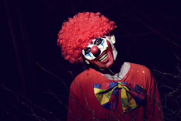 closeup of a scary evil clown wearing a red wig and a dirty costume, in the woods at night