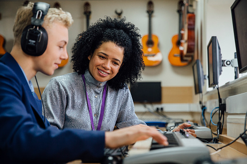 Female teacher is sitting with one of her students in a music lesson at school. He is learning to play the keyboard and is wearing headphones.