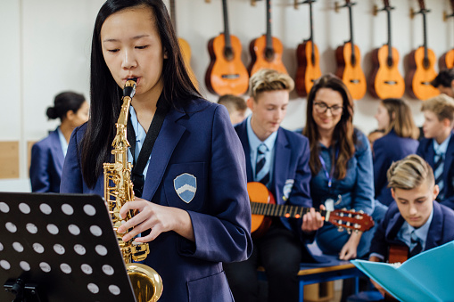 Teen student is playing the saxophone in her school music lesson. The rest of the class are in the background, out of focus.