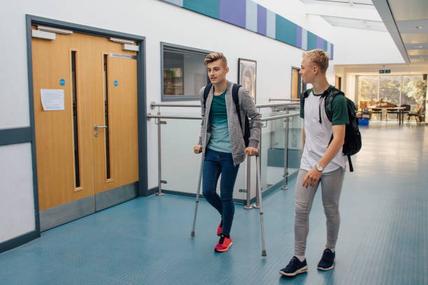 Accompanying Friend At School Teen students are walking down the school hall together. They have left lesson early, as one of the boys is on crutches and requires assistance from his friend. calm before the storm photos stock pictures, royalty-free photos & images