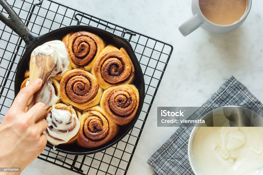 Woman's Hand Spreading Frosting Across Cinnamon Rolls in Skillet An anonymous woman's hand spreads cream cheese frosting on top of freshly baked cinnamon rolls. The rolls were baked in a cast iron skillet and there is a mug of hot coffee or tea and a bowl of frosting on the side. Cinnamon Bun Stock Photo