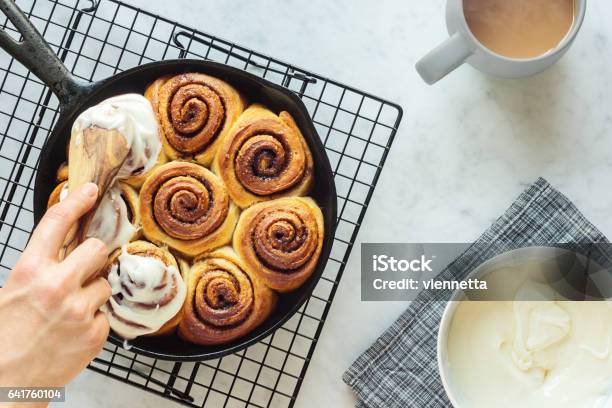 Womans Hand Spreading Frosting Across Cinnamon Rolls In Skillet Stock Photo - Download Image Now
