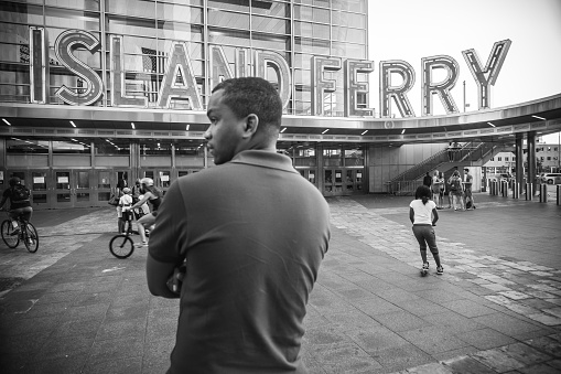 Lower Manhattan, New York City, USA. Young black man standing on the platoin front of the Island Ferry Terminal, looking to the side. Other people and kids riding bikes in the background. This is the place to get  The only non-vehicular Public transport connecting Manhattan with Norther borrows of NYC, Staten Island. This is a 5 miles, 25 minutes ride providing magnificent views of NYC harbour, The Statue of Liberty and more.