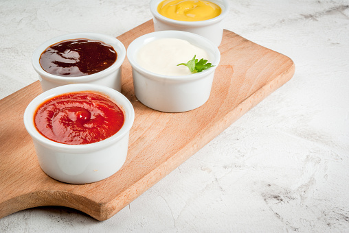 Classic set of sauces in white saucers: American yellow mustard, ketchup, barbecue sauce, mayonnaise. On cutting board white stone concrete table close view, copy space