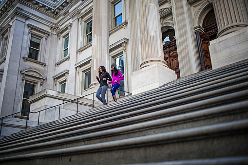 Low angle view of women jogging on steps. Full length of determined females are exercising outside building. Multi-ethnic friends are in sportswear.
